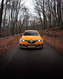 Une voiture de sport orange vibrante roulant sur une route forestière, entourée d'arbres d'automne.