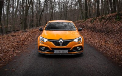 Une voiture de sport orange vibrante roulant sur une route forestière, entourée d'arbres d'automne.